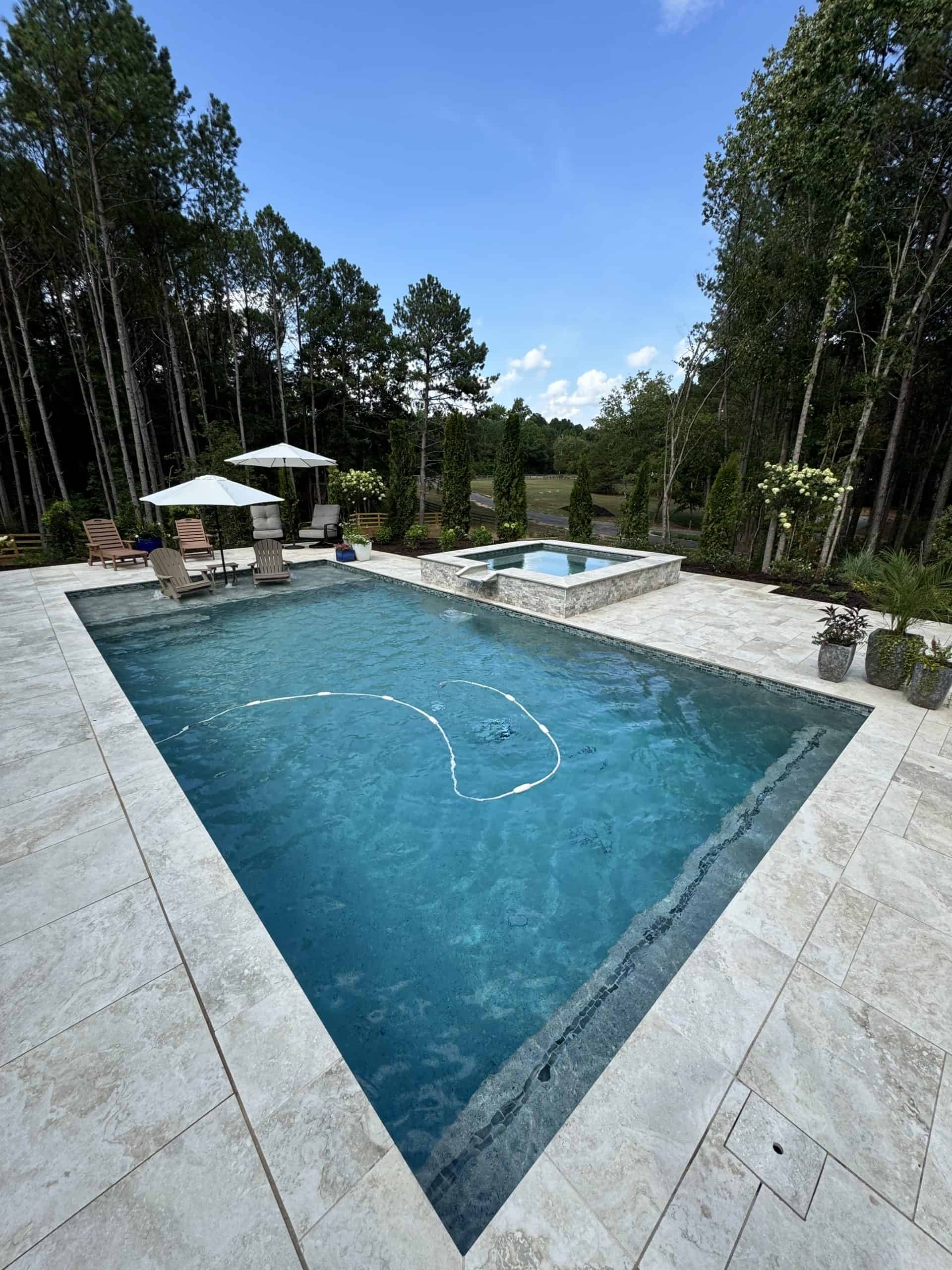 Luxury rectangular pool and elevated spa with travertine coping and deck, flanked by Italian cypress trees, ornamental container plantings, and pastoral field views at Burras Oak Lane in Cumming, Georgia, designed by Garden Atlantic