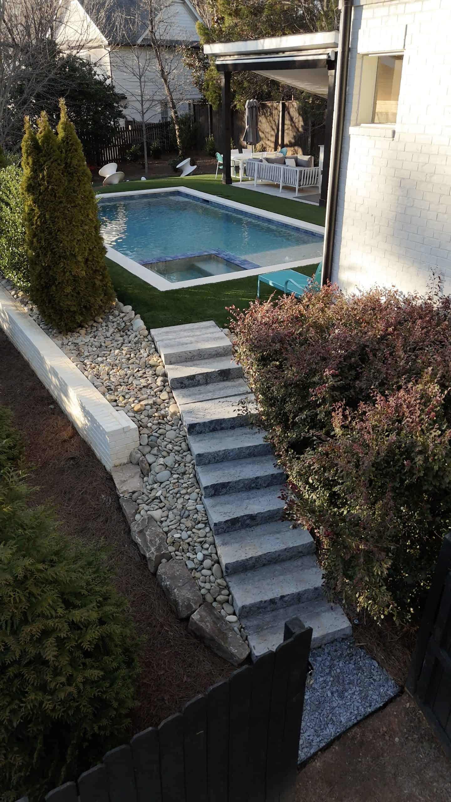 Side yard staircase with travertine steps and river rock dry creek bed leading to a rectangular pool with integrated spa, synthetic turf lawn, and covered outdoor living area at a Sandy Springs, Georgia residence designed by Garden Atlantic