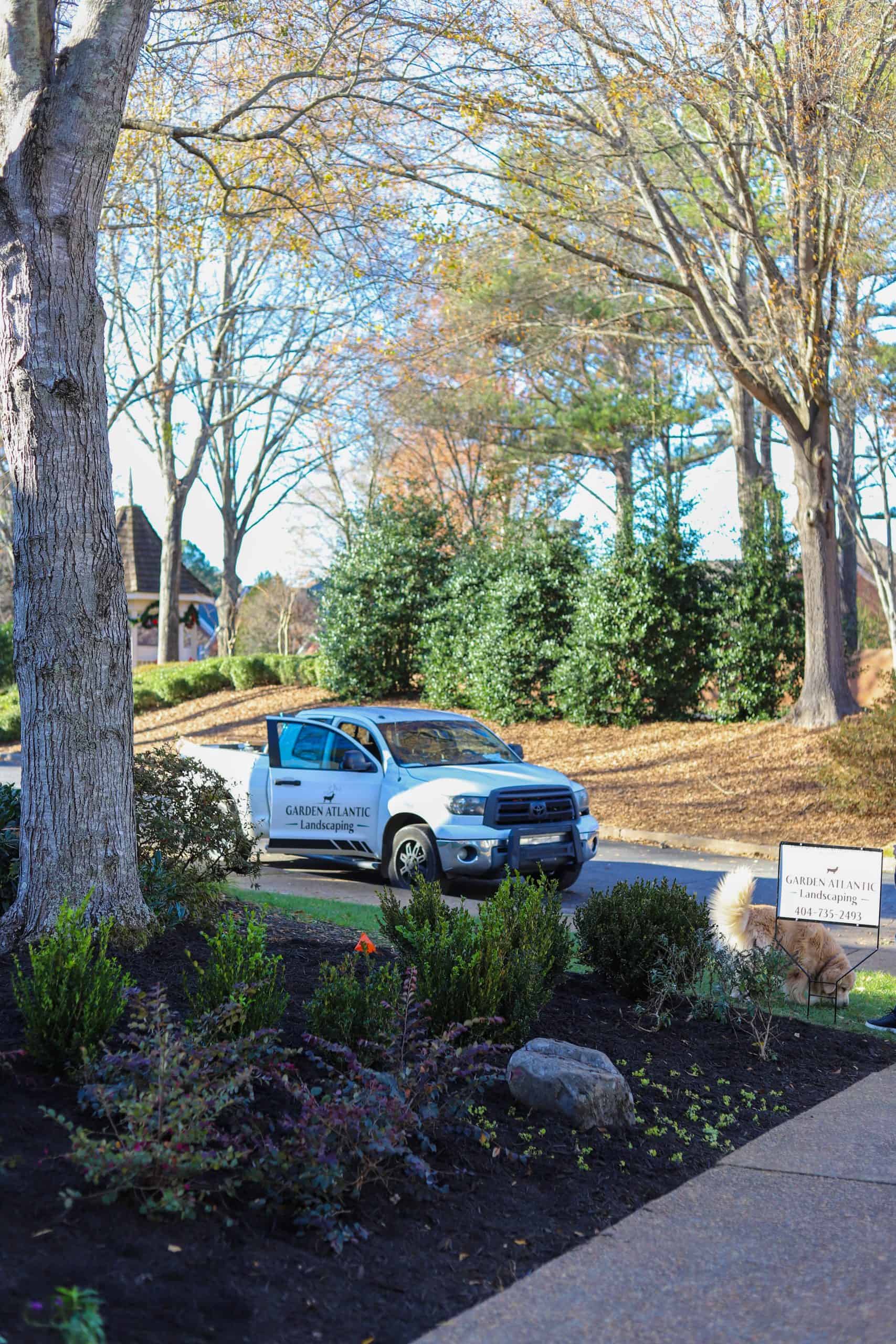 Wide view of Summerour Vale estate entry planting with Garden Atlantic truck and yard sign in foreground, featuring perennial beds, barberry, natural boulders, and mature tree canopy in Atlanta neighborhood
