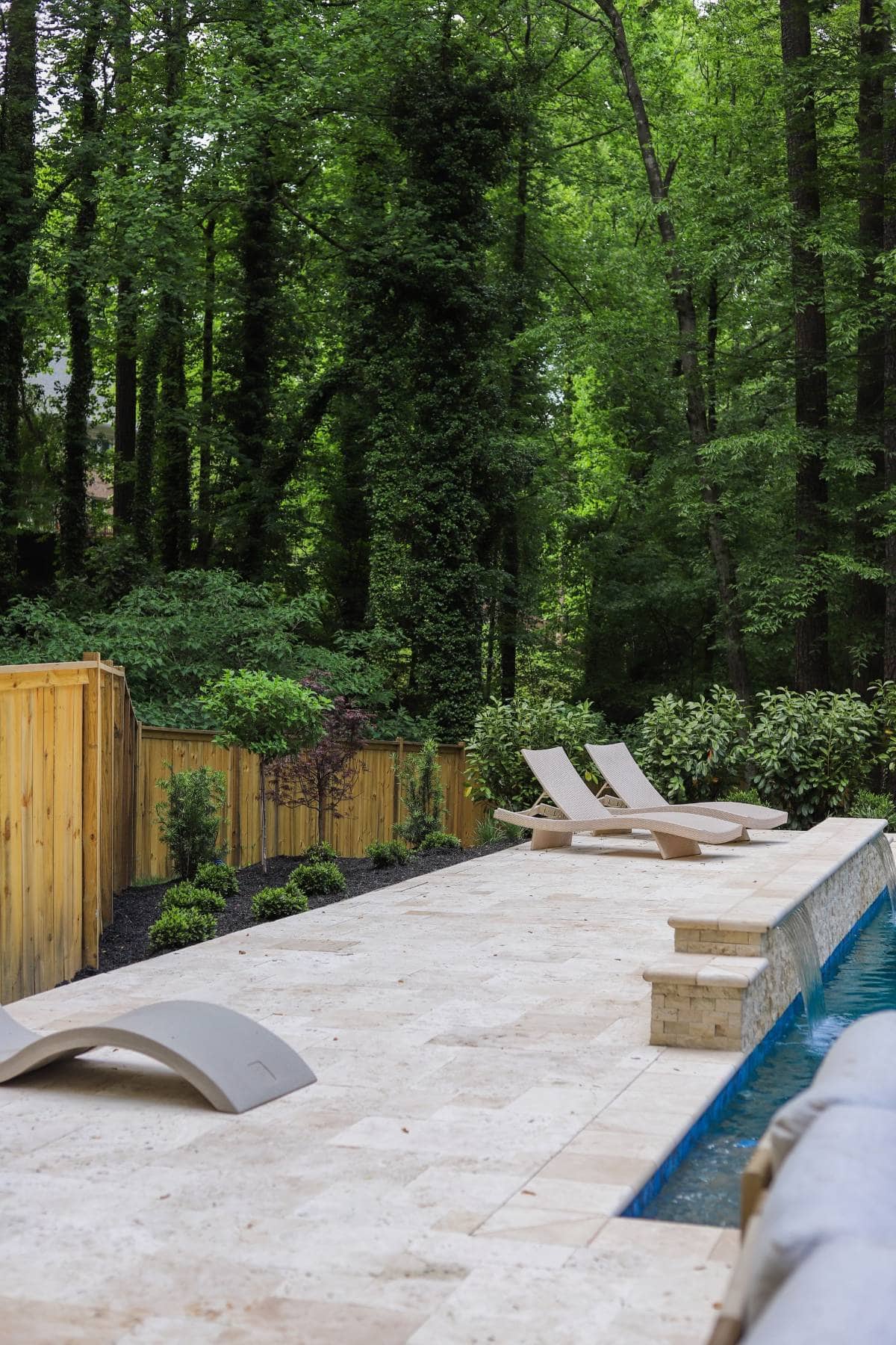 Travertine pool deck with modern loungers, stacked stone water feature, and privacy planting at Tynecastle, Atlanta by Garden Atlantic